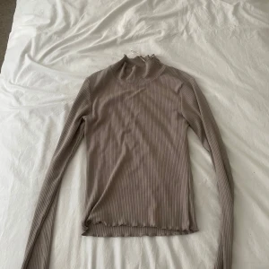 Beige ribbad polotröja - Supersnygg beige ribbad polotröja med lång ärm och lätt vågig kant vid hals och nederdel. Perfekt att styla med jeans eller kjol för en clean look. Tunn och stretchig, passar både lager på lager eller som den är.