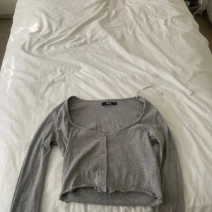 Grå croppad kofta från Bik Bok - Säljer en grå croppad kofta från Bik Bok med lång ärm och knappar framtill. Tröjan har en djup v-ringning och är tillverkad i mjuk, ribbad bomullsblandning. Perfekt att styla med höga jeans eller kjol.