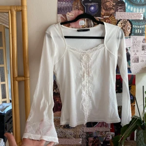 Vit spetsblus från KappAhl - Supersöt vit blus från KappAhl med långa ärmar och romantiska spetsdetaljer längs urringningen och framtill. Blusen har snörning framtill och lätt utsvängda ärmslut. Perfekt för dig som gillar en feminin och vintageinspirerad stil.