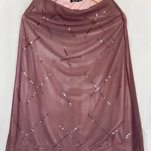 Rosa/Lila paljettkjol från Sisters - Snygg maxikjol från Sisters med glittriga paljettdetaljer i korsmönster. Klänningen är 75 cm lång och har ett tunt, transparent yttertyg över ett ljusare foder. Perfekt för dig som vill sticka ut med en unik look.