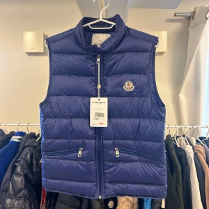 Blå moncler Gui Down väst. Storlek 1-S  - Skick 9/10 nypris 9300kr Snygg blå dunväst från Moncler med klassisk logga på bröstet. Västen har två dragkedjefickor framtill och stängs med dragkedja. Den är quiltad och har en hög krage för extra värme. Perfekt för lager-på-lager och streetstyle. 