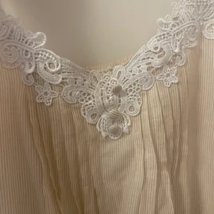 Beige linnetopp med spets från Atmosphere - Supersöt beige linnetopp med tunna axelband och vita spetsdetaljer längs både ringning och nederkant. Tunt, randigt tyg som ger en luftig känsla. Perfekt för dig som gillar romantiska och feminina vibes.