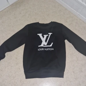 Svart Louis Vuitton sweatshirt - Svart sweatshirt från Louis Vuitton med stor vit LV-logga och text på bröstet. Tröjan har rund halsringning och långa ärmar. Perfekt för dig som gillar stilrena och exklusiva plagg med tydlig logga.
