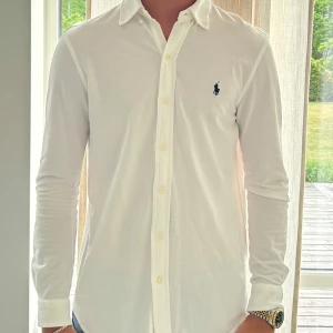 Vit skjorta från Polo Ralph Lauren - Klassisk vit skjorta från Polo Ralph Lauren i lätt mesh-bomull. Skjortan har lång ärm, knappar framtill och den ikoniska broderade loggan på bröstet. Perfekt passform och stilren look som funkar till jeans eller chinos.
