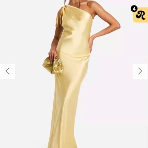 Champagnefärgad långklänning i satin - Elegant långklänning i champagnefärgad satin med oneshoulder och knyt-detalj på axeln. Klänningen har en slank siluett och skimrande finish som ger en lyxig känsla. Perfekt för dig som vill sticka ut på bal eller fest. Aldrig använd med lappen kvar!