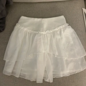 Vit volangkjol i chiffongstil - Supersöt vit kortkjol med dubbla volanger och flowig känsla. Kjolen har en hög midja och är gjord i ett tunt, lätt material som ger en drömmig look. Perfekt för dig som gillar romantisk och feminin stil.