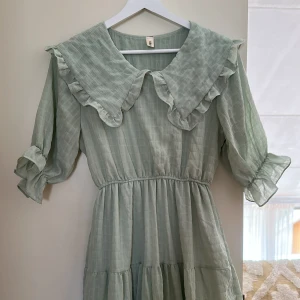 Ljusgrön långklänning med volangkrage - Superfin ljusgrön långklänning med volangkrage och trekvartsärmar. Klänningen har ett diskret rutmönster, markerad midja och volangdetaljer vid ärmslut och krage. Perfekt för dig som gillar romantisk och trendig stil.