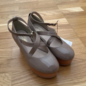 Beige platå ballerinaskor från NICE CLAUP - Snygga beige ballerinaskor med glansig finish och bred platåsula i trälook. Skorna har rund tå, korsade remmar över foten och justerbart spänne vid ankeln. Och kommer med extra band. Perfekt för dig som vill sticka ut med en unik och trendig look.