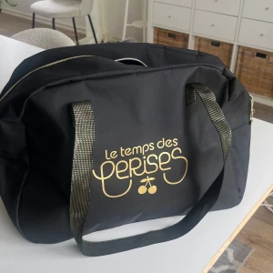 Svart weekendbag Le Temps des Cerises - Stor svart weekendbag från Le Temps des Cerises med guldiga detaljer och logga framtill. Väskan har breda axelband, dragkedja med körsbärsdetalj och är tillverkad i slitstark polyester. Perfekt för gymmet 