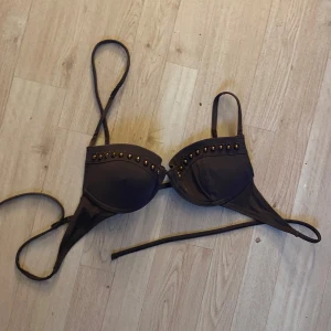 Svart bikiniöverdel med nitar - Snygg svart bikiniöverdel med coola nitar längs kanten på kuporna. Bikinin har vadderade kupor och smala axelband samt knyt i ryggen för perfekt passform. Perfekt för stranden eller poolen!
