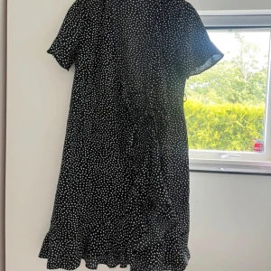 Svart prickig kortklänning med volang - Svart kortklänning med vita prickar och volangkant nertill. Klänningen har korta ärmar och knyt i midjan för en snygg passform. Perfekt för dig som gillar en lekfull och trendig stil. Passar XS/S