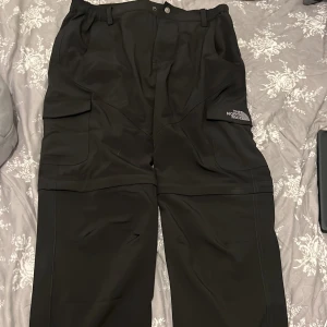 Svarta täckbyxor från The North Face - Svarta täckbyxor från The North Face med stora cargofickor på sidorna och broderad logga. Byxorna har avtagbara ben med dragkedja så de kan bli shorts. Perfekta för vinterns äventyr och outdoor-aktiviteter.