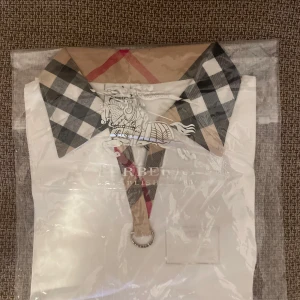 Vit Burberry skjorta med rutig krage - Snygg vit skjorta från Burberry med klassisk rutig krage och matchande detalj vid halsen. Skjortan har en stilren design med tydlig Burberry-tag och cool metallring framtill. Perfekt för dig som vill sticka ut med en ikonisk look.