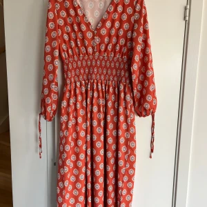 Roströd långklänning med blommigt mönster - Snygg roströd långklänning med vitt och blått blommigt mönster. Klänningen har v-ringning, smock i midjan och långa ärmar med knyt vid ärmslut. Luftigt material och flowy passform, perfekt för varma dagar.