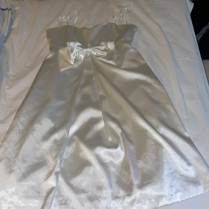 Vit satin klänning med rosett från Savann - Superfin vit kortklänning i satin från Savann med smala axelband och en stor rosett framtill. Klänningen har en glansig finish och en enkel, elegant design som verkligen sticker ut. Perfekt för dig som vill ha en chic och stilren look.