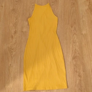 Gul ribbad midiklänning med snörning - Snygg gul ribbad klänning med tight passform och midilängd. Klänningen har hög hals, är ärmlös och har en öppen rygg med snörningsdetalj som ger en cool look. 