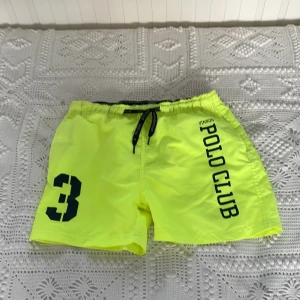 Neongula badshorts Vinson Polo Club - Neongula badshorts från Vinson Polo Club med svart snörning i midjan och tryckta detaljer som stort nummer 3 och logga på ena benet. Shortsen har meshfoder och elastisk midja för skön passform. Perfekta för stranden eller poolen!