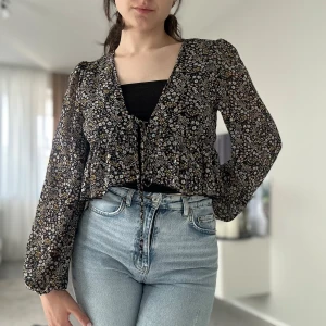 Blommig svart blus med snörning - Supersöt blommig blus i svart med små vita och gula blommor. Lätt transparent material, långa puffiga ärmar och v-ringning. Knytband framtill och volangkant nedtill ger en romantisk vibe. Perfekt att styla med jeans för en chill look.