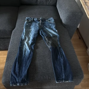 Dsquared2 blå jeans med patch - Säljer ett par blå Dsquared2 jeans med coola slitningar och en svart patch på ena benet. Jeansen har knappar med logga, klassisk femficksmodell och snygg tvätt. Perfekt för dig som gillar streetstyle och unika detaljer.