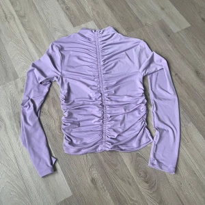 Ljuslila långärmad topp från Gina Tricot - Supersnygg ljuslila långärmad topp från Gina Tricot med rynkade detaljer längs hela fram- och baksida. Stretchigt och mjukt material som sitter tajt och ger en cool look. Perfekt att styla med jeans eller kjol för en trendig vibe.