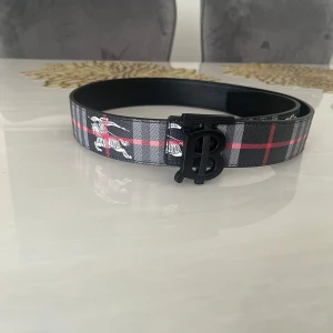 Burberry rutig bälte med B-spänne - Snyggt bälte från Burberry med klassiskt rutigt mönster i svart, grått, vitt och rött. Bältet har ett svart B-formad metallspänne och detaljer med Burberry-ryttare. Perfekt accessoar för att lyfta din outfit.