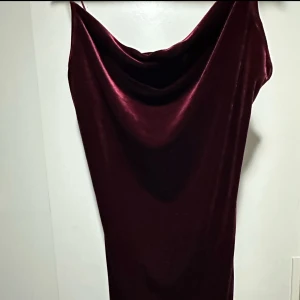 Vinröd sammetsklänning med draperad urringning - Säljer en kortklänning i vinröd sammet med smala axelband och snygg draperad urringning. Klänningen har en elegant, figurnära passform och är perfekt för dig som vill sticka ut med en lyxig vibe. Materialet är mjukt och skimrande, vilket ger en exklusiv känsla.