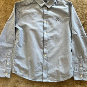 Ljusblå skjorta från Henri Lloyd - Klassisk ljusblå skjorta från Henri Lloyd med broderad logga på bröstet. Skjortan har lång ärm, knappar framtill och en snygg krage. Tillverkad i mjuk bomull som känns skön mot huden. Perfekt för en clean och stilren look.