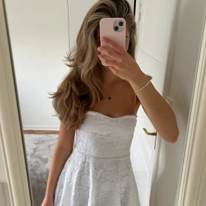 Vit axelbandslös spetsklänning - Superfin vit kort klänning med axelbandslös design och vacker spets över hela tyget. Klänningen har en figurnära överdel och utsvängd kjol, perfekt för sommarens alla tillfällen. Söt och fräsch look med romantiska vibbar. Perfekt för midsommar eller all white party!