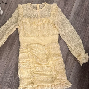 Gul spetsklänning med volanger - Superfin ljusgul kortklänning i spets med långa ärmar och volangdetaljer. Klänningen har rund halsringning, transparenta ärmar och dragkedja bak. Perfekt för dig som gillar romantisk och feminin stil. Perfekt för midsommar! Storlek S, endast använd vid en midsommar.