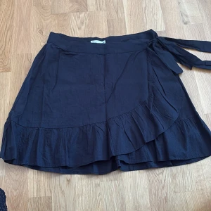 Mörkblå  omlottkjol från NLY Trend - Mörkblå omlottkjol från NLY Trend i storlek 38. Kjolen har volangkant nertill och knyts i midjan för en snygg passform. Perfekt för dig som gillar en enkel men trendig look.