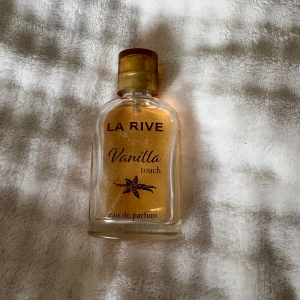 La Rive Vanilla Touch parfym - La Rive Vanilla Touch eau de parfum i en genomskinlig glasflaska med guldig kork. Parfymen har en varm, söt doft av vanilj och flaskan är dekorerad med en vaniljblomma på framsidan. Perfekt för dig som älskar mjuka och söta dofter.