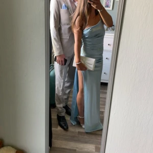 Ljusblå långklänning med slits - Supersnygg ljusblå långklänning i satinliknande material med en axel och hög slits på sidan. Klänningen har en tight passform och draperad effekt över bysten. Perfekt för bal eller student. Den är endast använd 1 gång och inga defekter förekommer. Den har även en dragkedja på sidan och axelbandet är justerbart😊