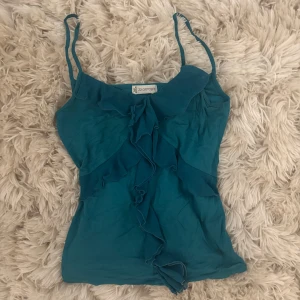 Turkos linnetopp med volang - Supersöt turkos linnetopp från 22 Octobre med smala axelband och volangdetaljer framtill. Snygg att styla med jeans eller kjol för en cool look.