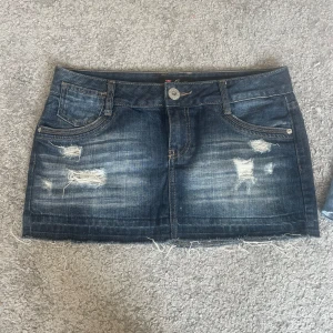 Lågmidjad jeanskjol från guess - Snygg mörkblå jeanskjol från Guess med slitna detaljer och fransig nederkant. Klassisk femficksmodell med dragkedja och knapp framtill. Perfekt för dig som gillar en edgy och avslappnad stil.