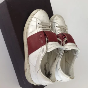 Valentino Open sneakers vit/röd skinn - Valentino Open sneakers i vitt skinn med bred röd skinnpanel över vristen. Klassisk rund tå, snörning och ikoniska nitar baktill på sulan. Perfekta för dig som gillar lyxiga och stilrena sneakers med en twist. Använt skick!