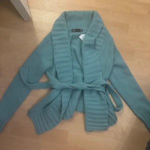 Ljusblå stickad kofta från Zara - Mysig ljusblå stickad kofta från Zara med bred ribbad krage och knytskärp i midjan. Perfekt för lager-på-lager och chill dagar. Långärmad och med snygga ribbade detaljer vid ärmslut och nederkant.