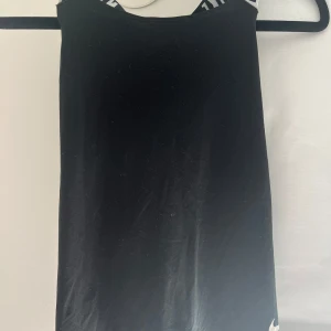 Svart Nike linnetopp med logoband - Svart linnetopp från Nike med vita, korsade axelband där det står NIKE i svart. Toppen är i Dri-FIT-material som andas och har en vit Nike-logga nertill. Perfekt för träning och sportiga outfits. Linnet är i barnstrorlek L/146-156! 🏆💃