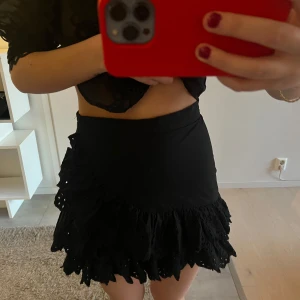 Svart volangkjol med broderade detaljer - Supersöt svart kortkjol med volanger och broderade håldetaljer! Perfekt vid flera tillfällen!🥰🥰de är en dragkedja i midjan! Den är i M men rätt liten i storleken 