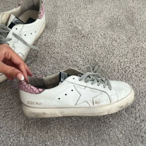Golden Goose vita sneakers med rosa detalj - Golden Goose sneakers i vitt skinn med grå snörning och ikonisk stjärna på sidan. Baktill finns en rosa detalj med skalmönster som ger en unik touch. Platt sula och GGDB/MAY-tryck i guld på sidan. Perfekta för en cool och avslappnad stil.