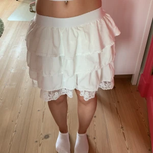 Vit volangkjol med spetsdetaljer - Supersöt vit kortkjol med flera volanger och bred resår i midjan. Kjolen har fina spetsdetaljer längst ner och broderade blommor i spetsen. Perfekt för dig som gillar en romantisk och lekfull stil. Kjolen har några lösa trådar. 