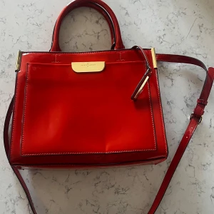 Röd handväska från Jasper Conran - Snygg röd handväska i glansigt material med guldiga metalldetaljer och både handtag och axelrem. Väskan har flera fack, varav ett med dragkedja, och stilren design med fyrkantig form. Väskan har fått ett fåtal skavanker från att ha legat oanvänd i en låda som syns på bilderna 3,4 och 5. Hör gärna av vid frågor eller funderingar.