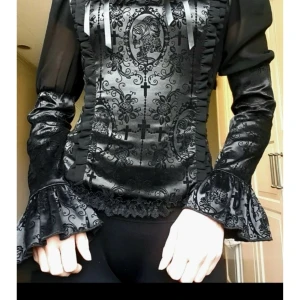 Blus Banned Apparel  - Blus från Banned Apparel.  Svart satin, detaljerad framsida med kors och porträttmotiv, samt volanger och spetsdetaljer runt halsringningen och nederkanten.  Ärmarna med kombination av puffärmar i schiffong och åtsittande partier med spets. Baksidan har en korsettsnörning som ger en justerbar passform och dold dragkedja.   Storlek XS  Använd vid ett tilfälle, nyskick!   Se gärna mina andra amnonser, samfraktar!