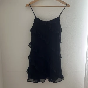 Svart volangklänning Gina Tricot - Supersöt svart kortklänning från Gina Tricot med tunna axelband och flera volangdetaljer framtill. Lätt och luftig känsla, perfekt för dig som gillar en feminin och trendig stil. Passar lika bra till sneakers som till klackar.