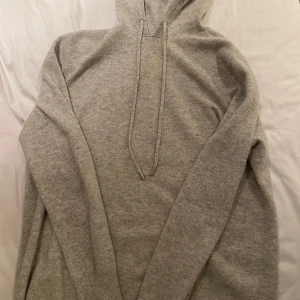 Ljusgrå stickad hoodie med huva - Mysig ljusgrå stickad hoodie med huva och snörning. Tröjan har långa ärmar och ribbade muddar vid ärmslut och nederkant. Perfekt för dig som gillar en enkel och stilren look med en soft känsla. 