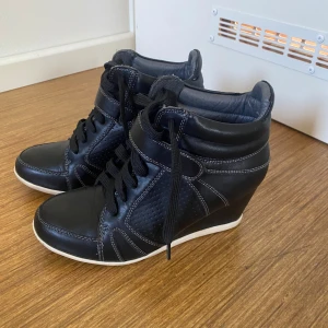 Svarta sneakers med kilklack, strl 37 - Svarta sneakers med vit sula och kilklack. Skorna har snörning, kardborreband och är gjorda i skinnimitation med perforerade detaljer på sidorna. Perfekt för dig som vill ha en sportig look med lite extra höjd.