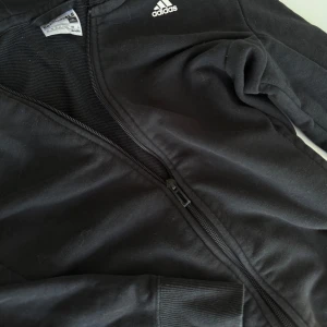 Svart Adidas hoodie med dragkedja - Svart hoodie från Adidas Essentials med vit logga på bröstet och stort Adidas-tryck längs ena ärmen. Jackan har huva, hel dragkedja och ribbade muddar. Tillverkad i mjuk bomullsmix. Använd några gånger. Ord pris 650kr, pris kan diskuteras.