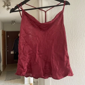 Satin topp med tunna axelband - Säljer en vinröd satin topp med tunna dubbla axelband och snygg vattenfalls urringning. Toppen har en lös passform och är tillverkad i ett glansigt material som ger en lyxig känsla. Perfekt för fina tillställningar. Kan strykas för att få bort veck.