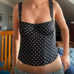 Svart prickig korsett-topp - Supersnygg svart korsett-topp med vita prickar och breda axelband. Toppen har markerade kupor och söt spetskant längs överkanten och nederkanten. Perfekt för att lyfta vilken outfit som helst med en retrovibe. ( köpte på vinted och lappen är bortklippt så vet ej märket) 💖💖