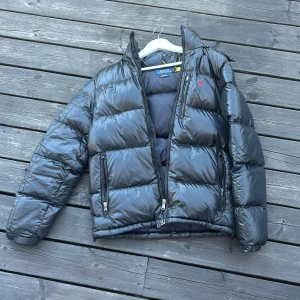 Svart pufferjacka från Polo Ralph Lauren - Snygg svart pufferjacka från Polo Ralph Lauren med klassisk röd logga på bröstet. Jackan har glansig finish, två stora fickor med dragkedja och hög krage. Perfekt för kalla dagar när du vill hålla stilen och värmen.