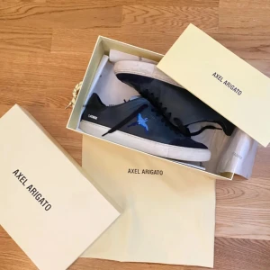 Axel Arigato svarta sneakers med blå fågel - Snygga svarta sneakers från Axel Arigato med broderad blå fågel på sidan och vit sula. Skorna har svarta snören och detaljer i skinn samt mocka vid tån. Perfekta för dig som gillar stilrena men unika sneakers. Har ett jack på sidan av skog som inte syns med dom på